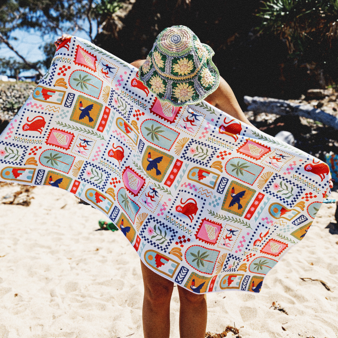 Lady holding a colorful patterned sand free towel wearing a hat on white sand beach