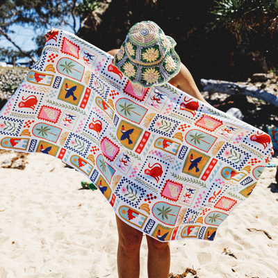 Lady holding a colorful patterned sand free towel wearing a hat on white sand beach