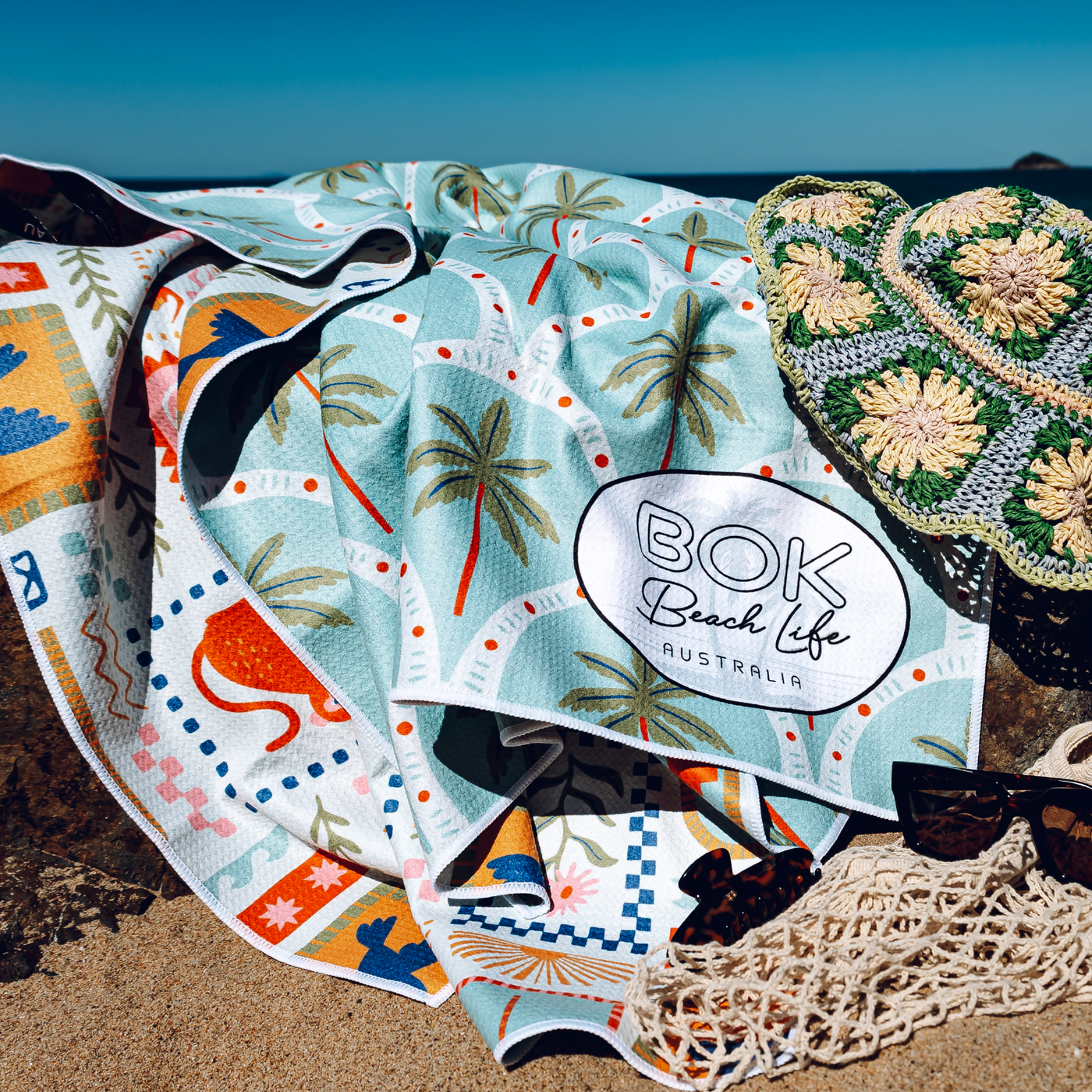 Colorful beach towel with palm tree design and 'BOK Beach Life Australia' branding on a sandy background.