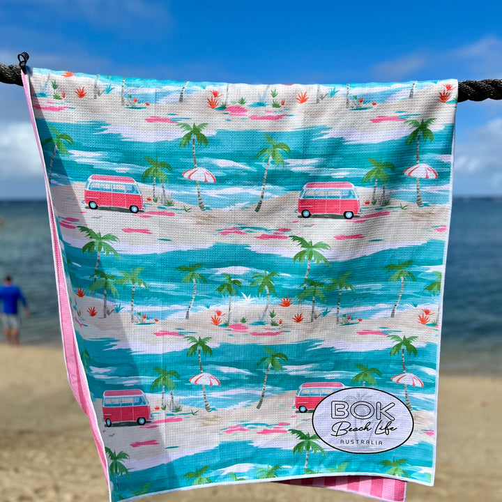 Beach towel with a tropical design featuring palm trees, beach umbrellas, and vans, hanging on a rope with ocean and sky in the background.
