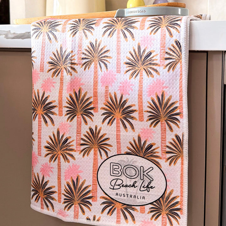 Kitchen towel with pink palm tree pattern and BOK Beach Life Australia logo on a kitchen counter.