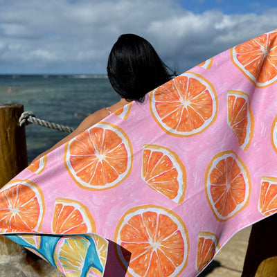 Woman holding a pink sand free beach towel featuring orange slices in the pattern she is standing by the ocean