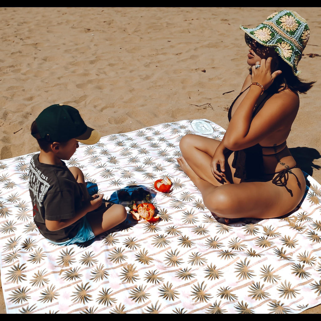 Woman and child sitting on the Swaying Palms Sand Free Beach Blanket by Bok Beach Life, relaxing and sharing snacks on the sand.