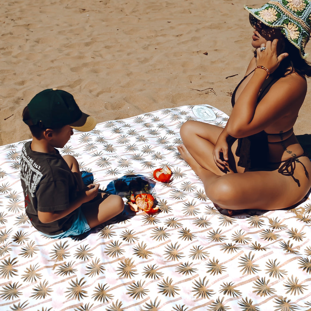 Woman and boy sitting on a tropical pink patterned sand free beachblanket at the beach with fruit.