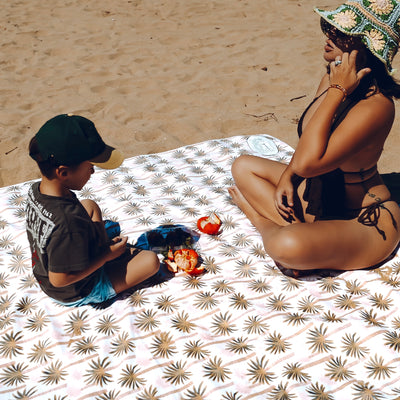 Woman and boy sitting on a tropical pink patterned sand free beachblanket at the beach with fruit.